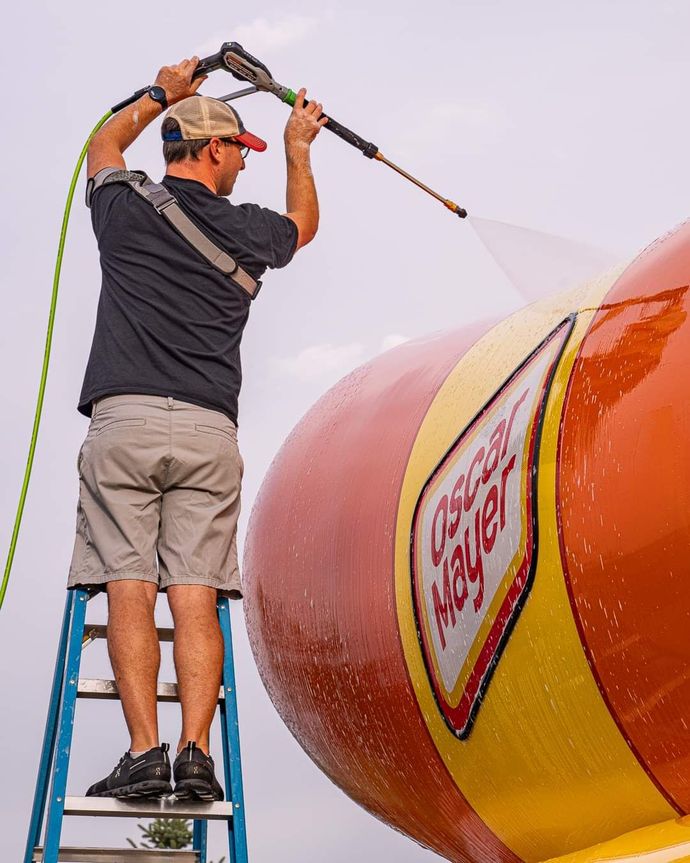 Worker on a ladder pressure-washing a giant orange and yellow buoy or tank with a hose.