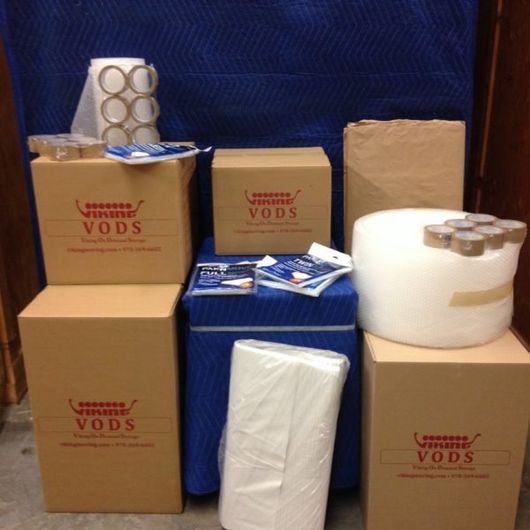 Stacked cardboard boxes and rolls of white packing material in a storage area, some labeled VODS.
