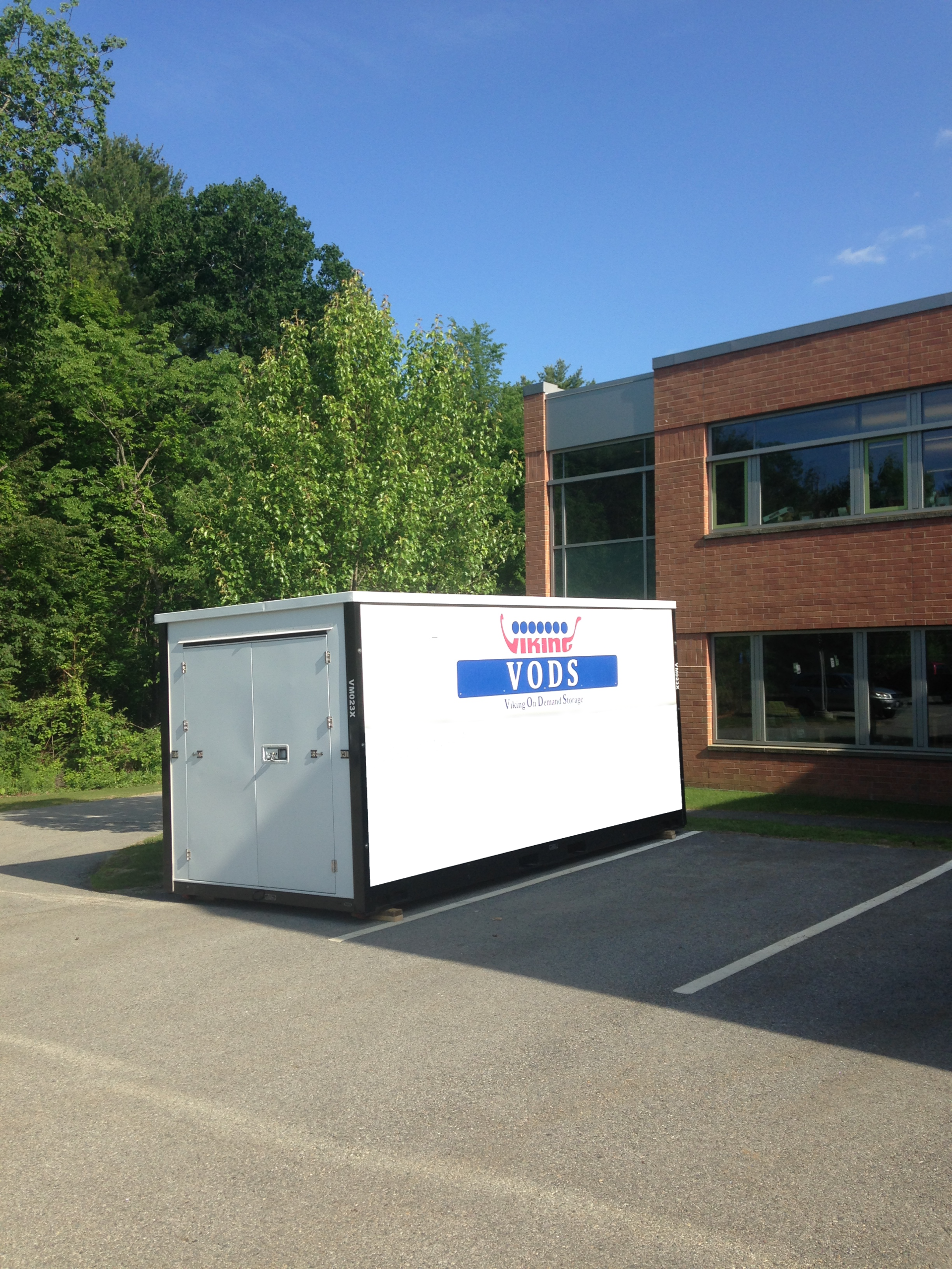White YDS dumpster in a parking lot beside a brick building and green trees under a blue sky