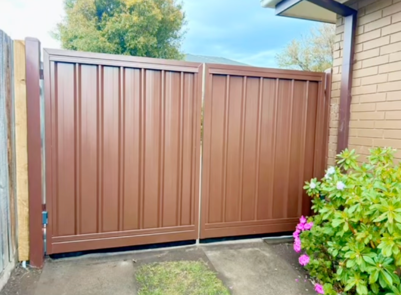 A brown gate is sitting next to a brick building.  — Talking Fences In Portarlington, VIC