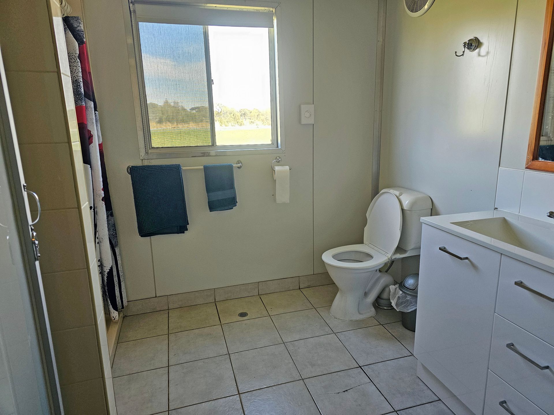 Small bathroom with toilet, sink cabinet, tiled floor, towels, and a window overlooking greenery