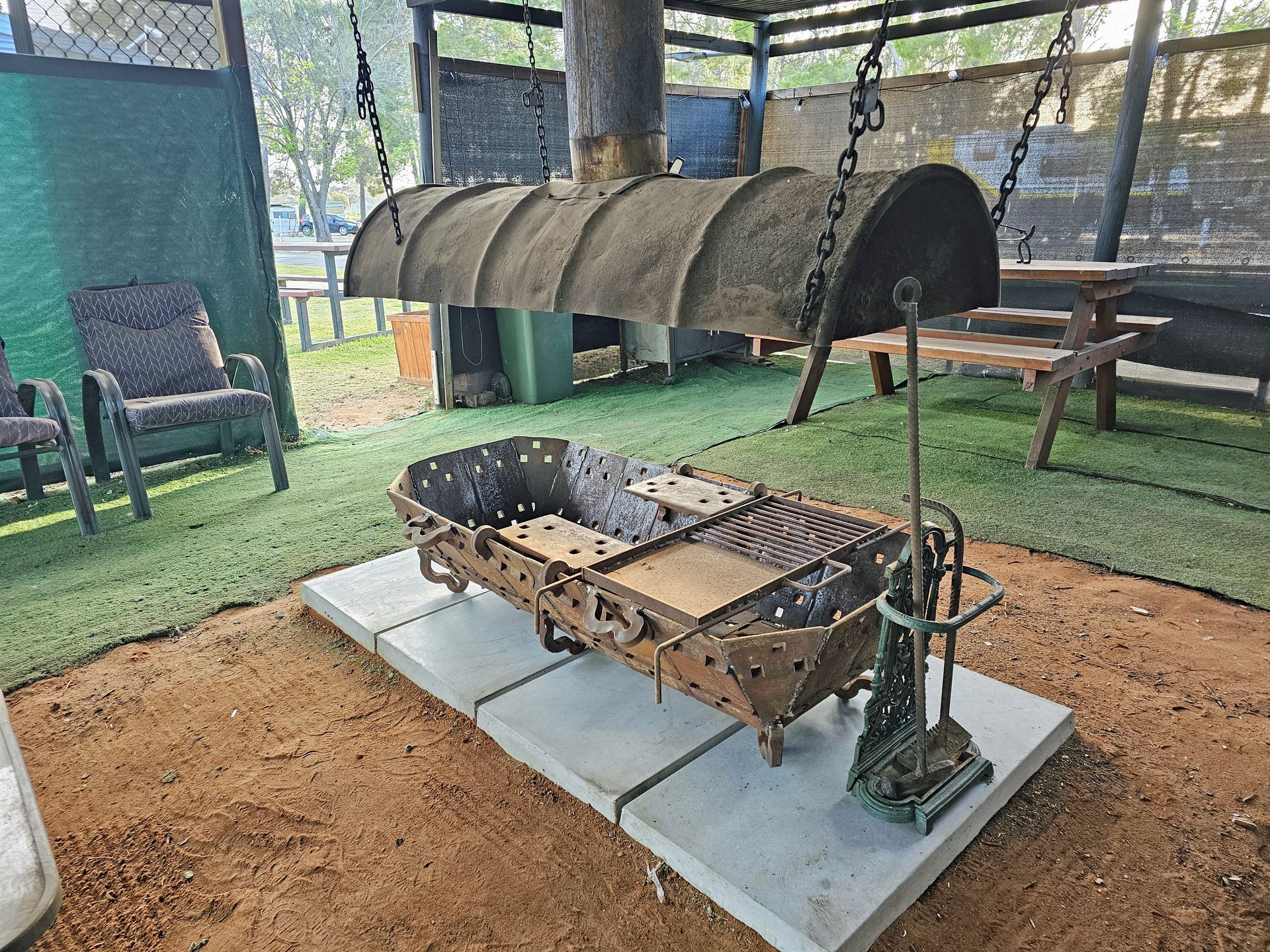Outdoor fire pit with a metal grill over a concrete pad, under a covered shelter with chairs nearby