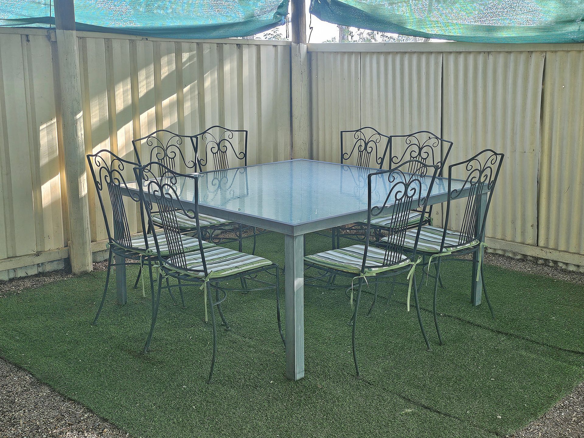 Outdoor patio table with four wrought-iron chairs on grass beside a beige fence and pool netting