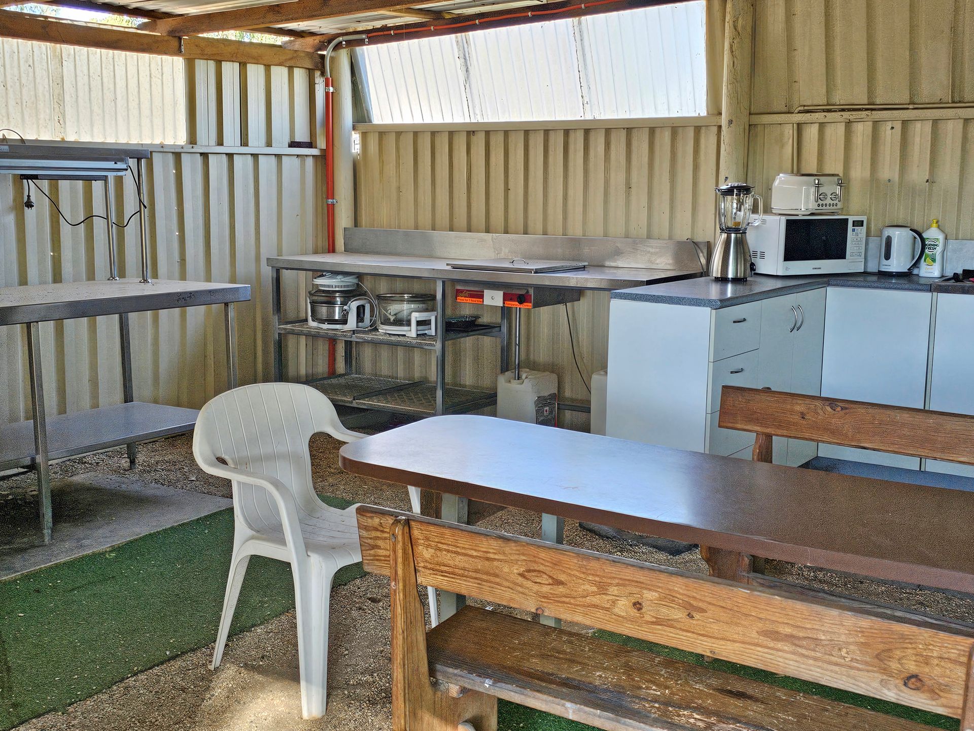 Simple kitchen with metal counters, white plastic chair, and wooden table inside a corrugated metal shelter