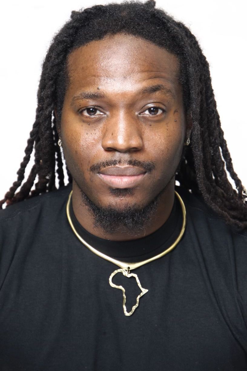 Man with long dreadlocks, wearing a black shirt and a gold necklace with an Africa-shaped pendant.