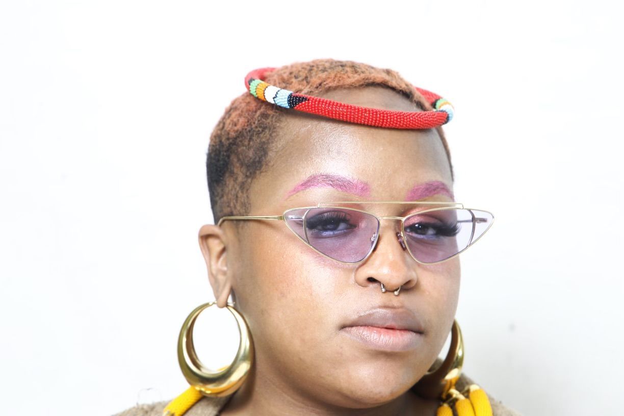 Person with short, dyed pink hair, wearing heart-shaped purple sunglasses, gold hoop earrings, a nose ring, and a red headpiece.