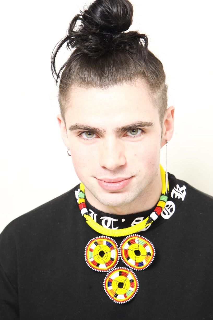 Man with topknot hairstyle wearing a beaded yellow necklace and black shirt.