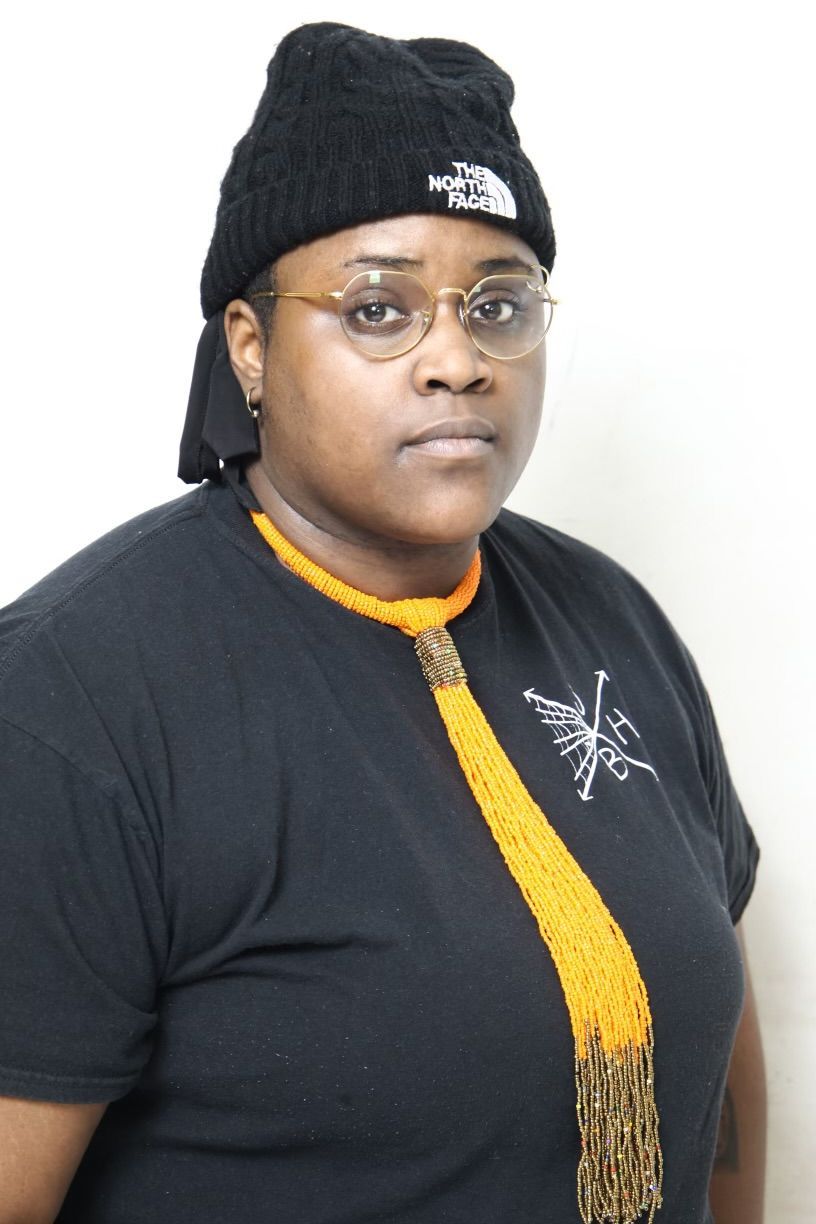 Person wearing glasses, black beanie, and orange beaded necklace, in front of a white wall.