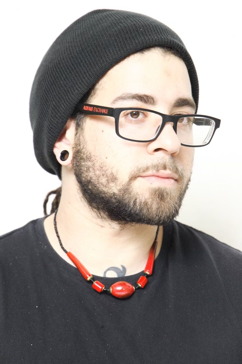 Man wearing glasses, black beanie, and red necklace. He has a beard and gauges.