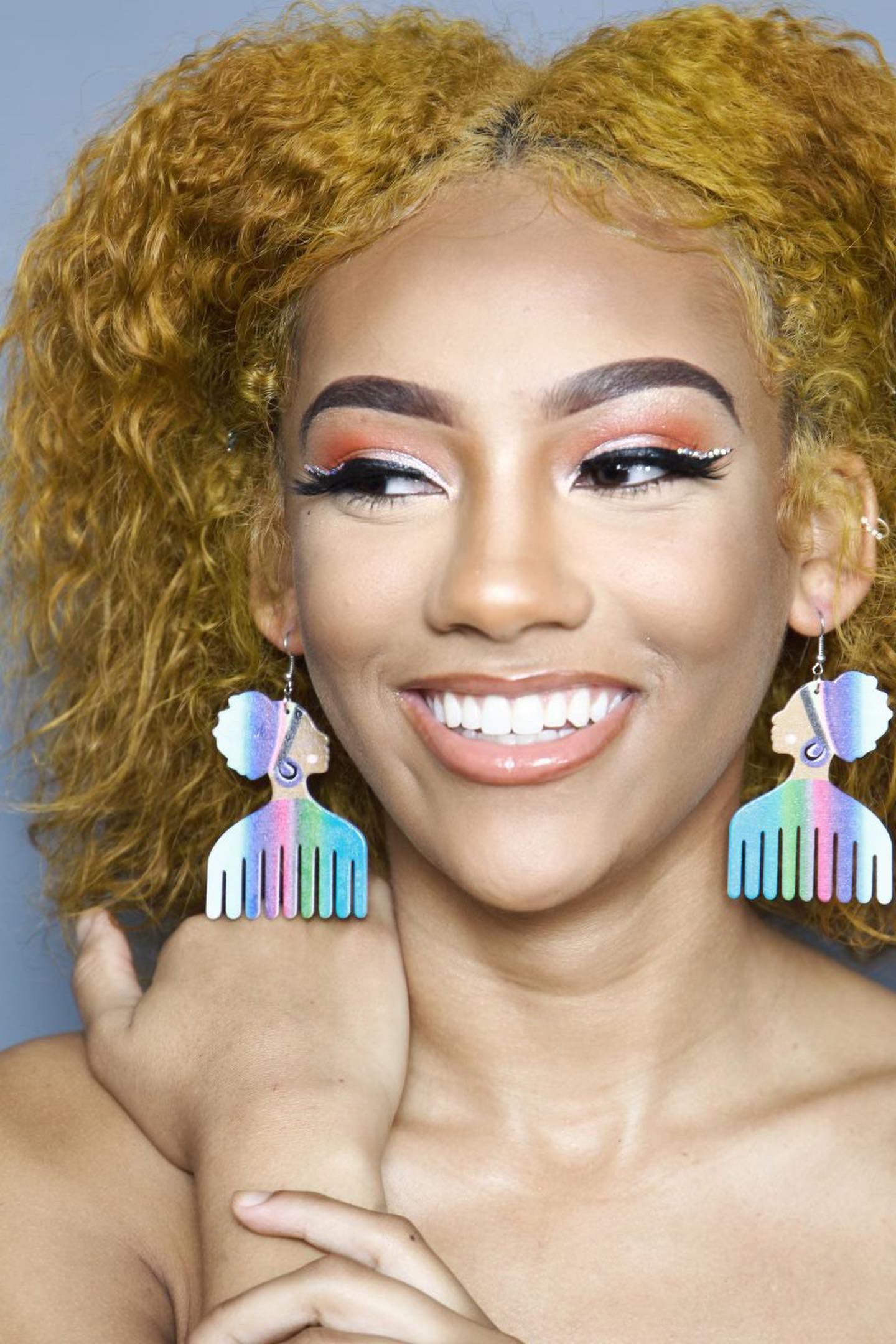 Woman smiling, wearing colorful comb earrings; light orange hair, vibrant eye makeup.