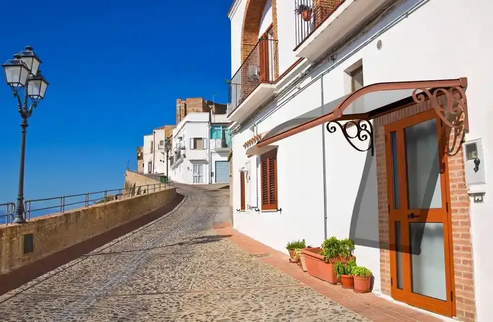 Una strada lastricata di ciottoli illuminata dal sole, affiancata da edifici bianchi e con vista sul mare.