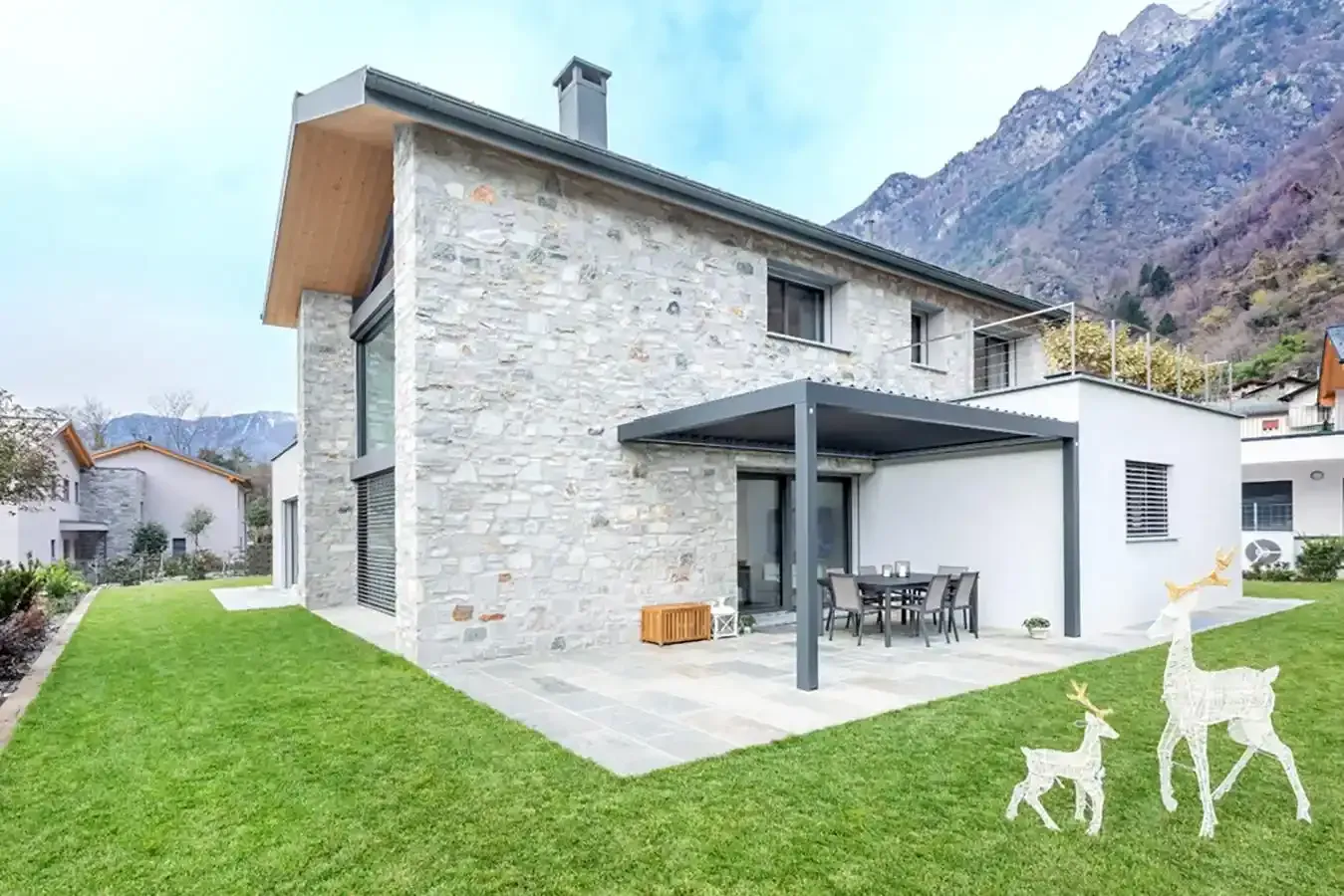 Casa in pietra con patio, prato verde e montagne sullo sfondo; due sculture di cervi bianchi nel cortile.