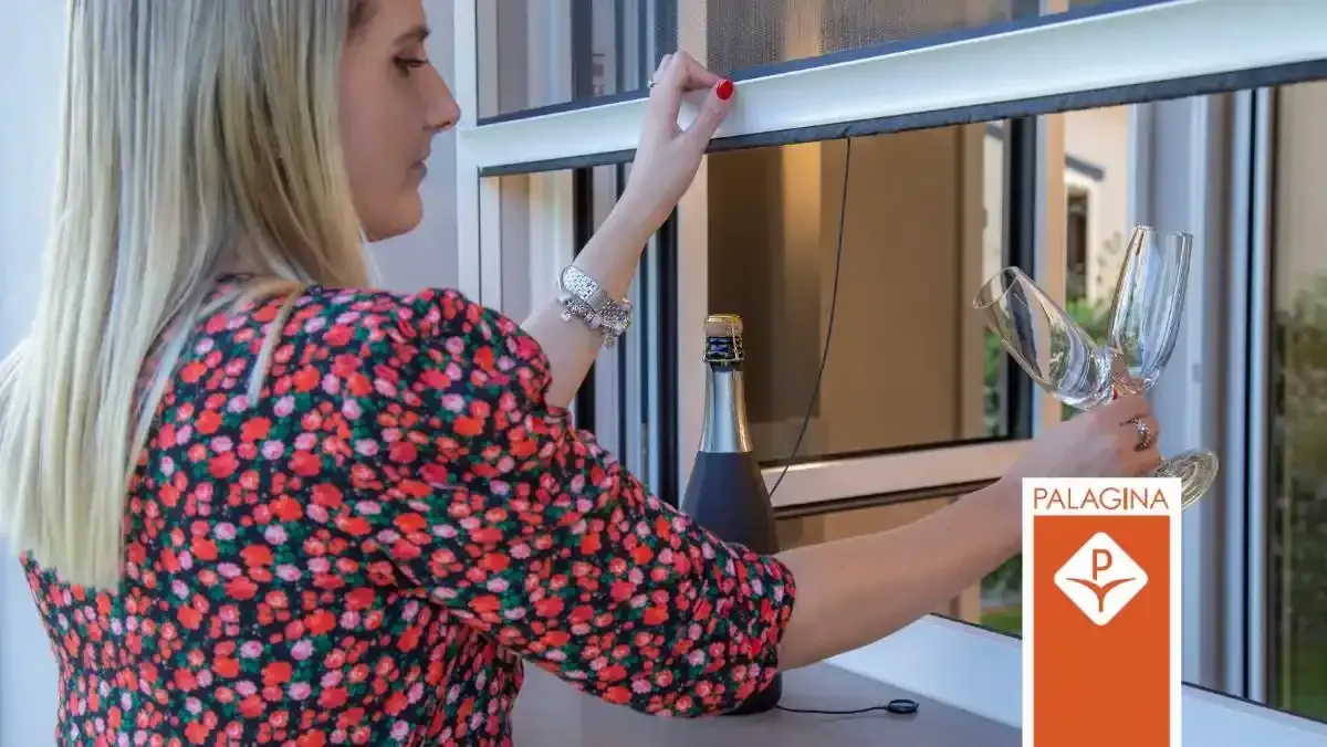Una donna con una blusa a fiori dipinge un telaio di una finestra con un pennellino accanto a una pianta in vaso.