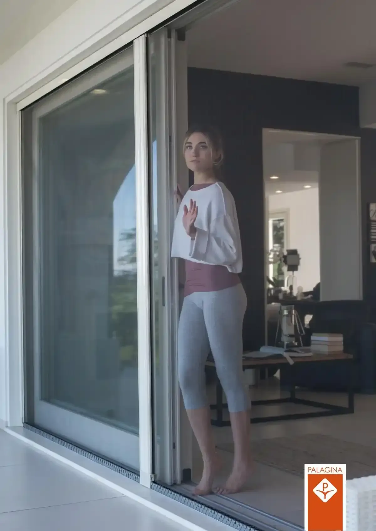 Persona in piedi su una porta scorrevole in vetro, con indosso una maglietta bianca e leggings grigi.