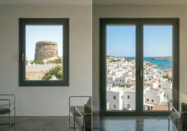 Camera moderna con due finestre che si affacciano su una cittadina costiera bianca, sul mare blu e su una torre di pietra all'esterno.