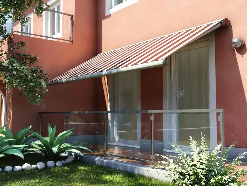 Patio con tenda da sole a righe, ringhiera in vetro, porte scorrevoli e piante rigogliose accanto a un edificio color pesca.