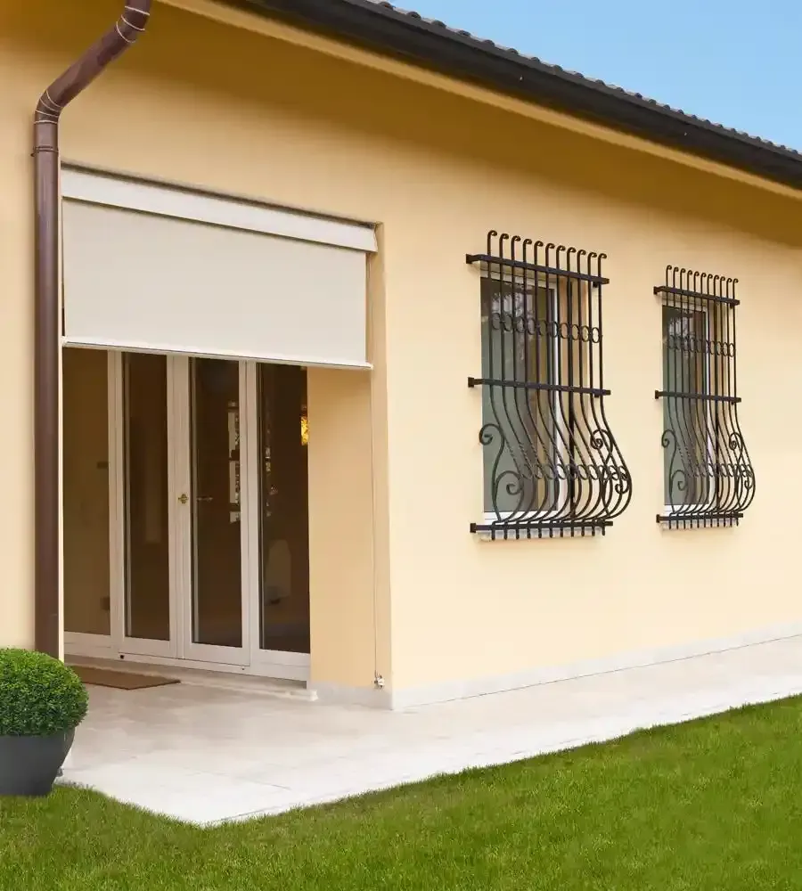 Esterno di una casa color crema con porte scorrevoli bianche, finestre con inferriate e un prato verde.