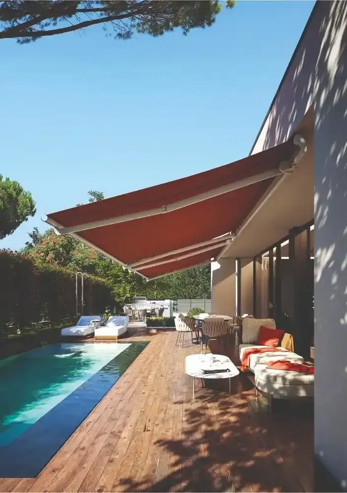 Patio moderno con tende da sole rosse accanto a una piscina, terrazza in legno e posti a sedere all'aperto in piena luce del sole.