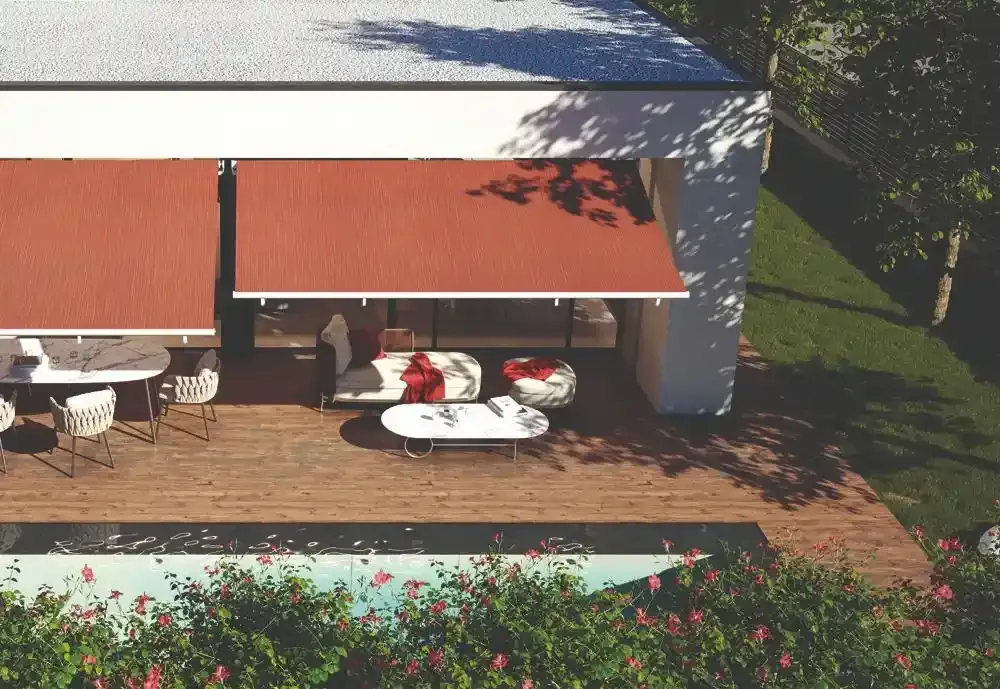 Patio esterno con tenda da sole arancione, tavolo e sedie bianchi e aiuola accanto alla casa.