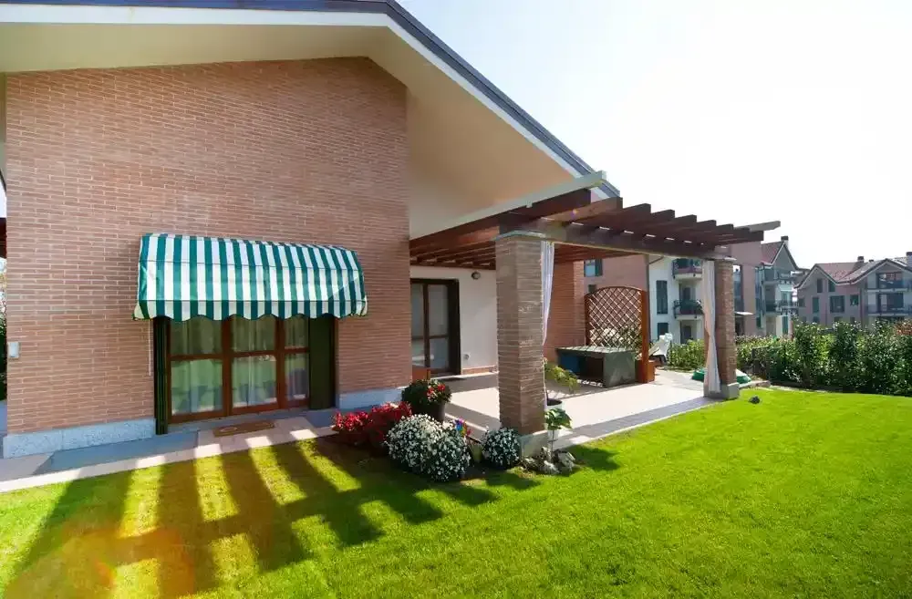 Casa in mattoni con tenda da sole a righe, patio coperto e prato verde brillante baciato dal sole