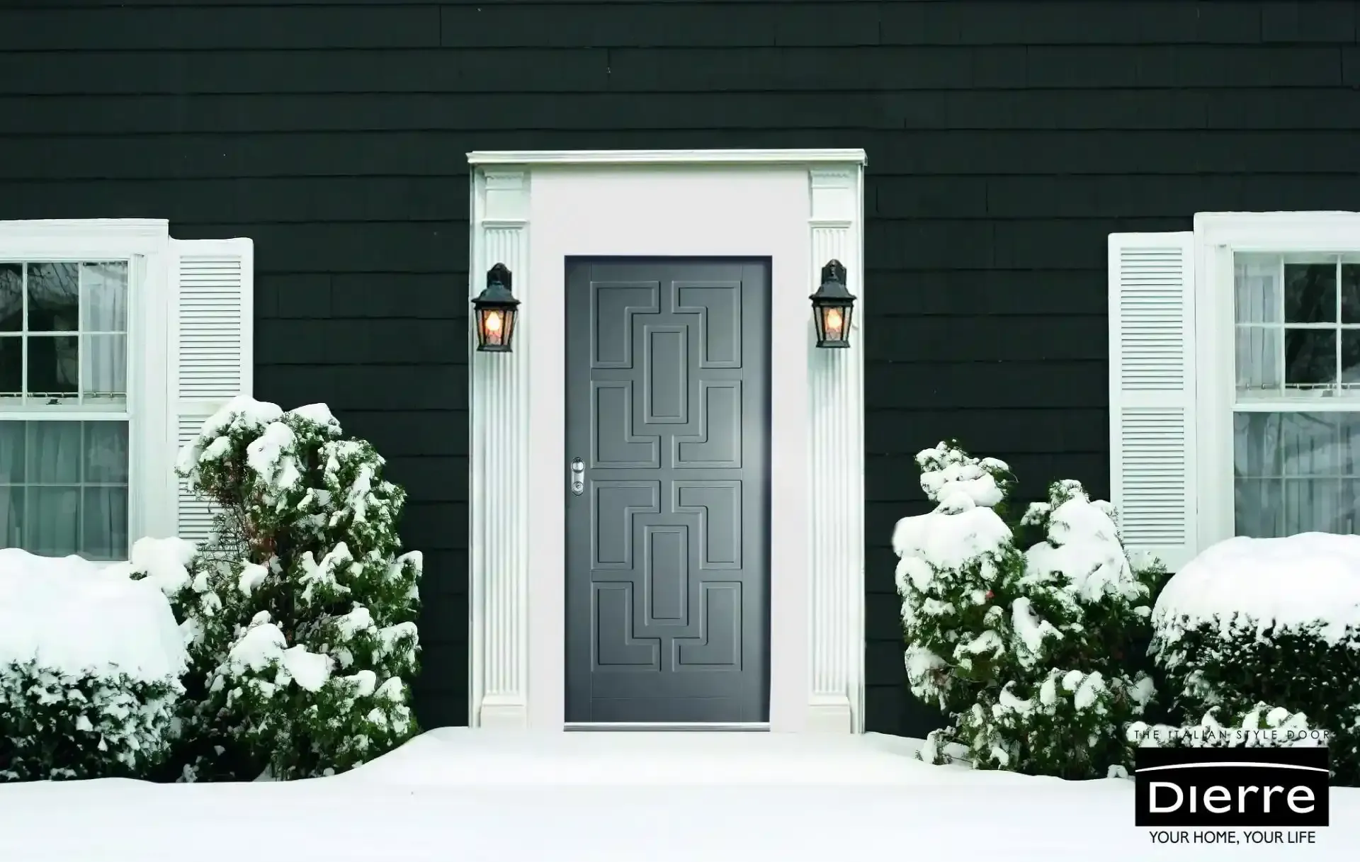 Porta d'ingresso innevata con cornici bianche, rivestimento esterno scuro e cespugli innevati su entrambi i lati.