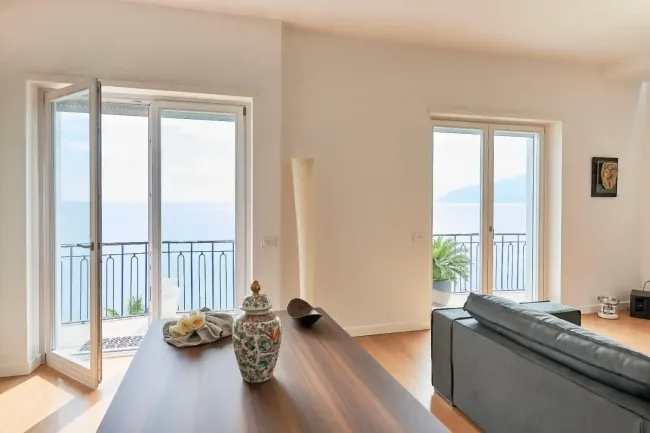 Luminoso soggiorno con porte finestre che si aprono sul balcone con vista sull'oceano, tavolo in legno e divano vicino a grandi finestre.