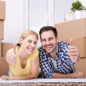 Smiling couple in a new home surrounded by moving boxes, giving thumbs up