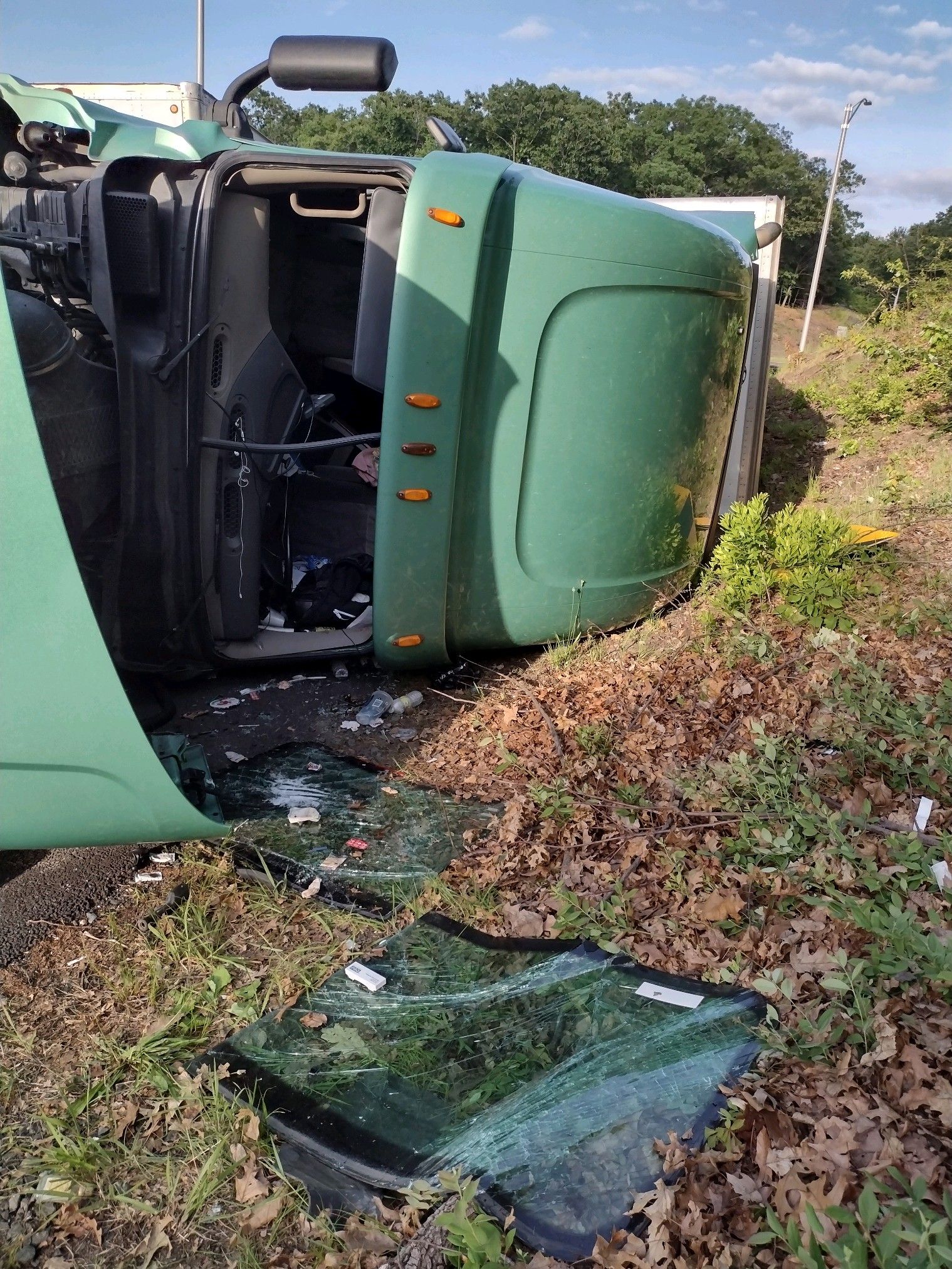 Green semi-truck on its side after accident; broken glass on the ground.