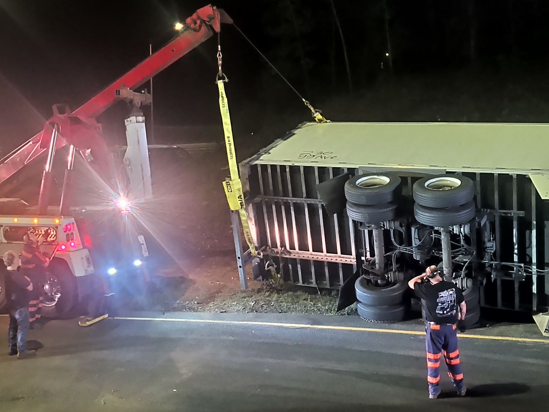 Tow truck uprighting a semi-trailer that has overturned on a highway at night.