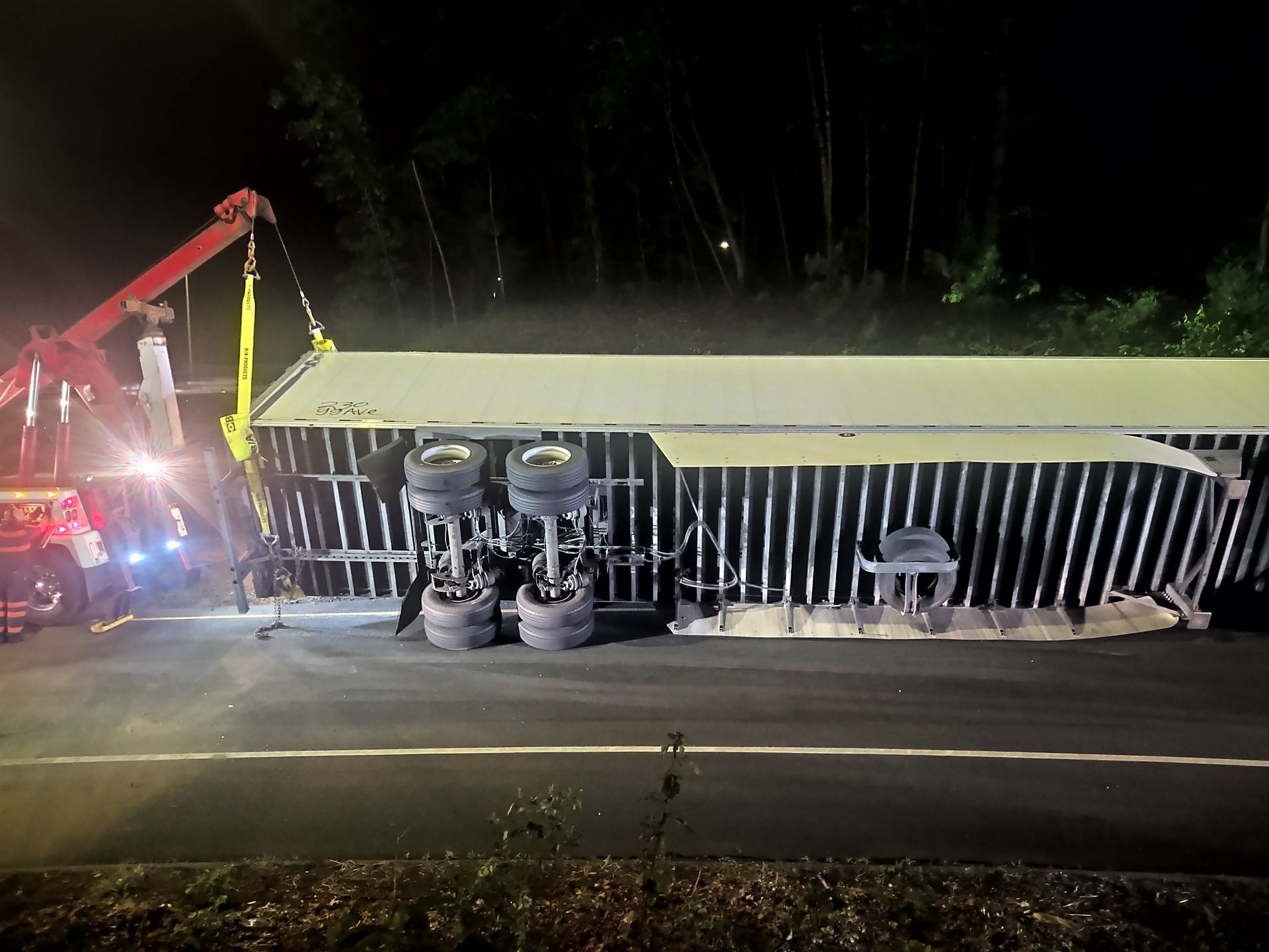 A tow truck lifts a semi-trailer lying on its side on a dark road.
