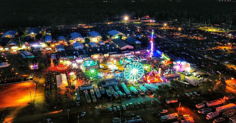 Aerial view of a brightly lit county fair at night, featuring a large Ferris wheel, carnival rides, and rows of campers.