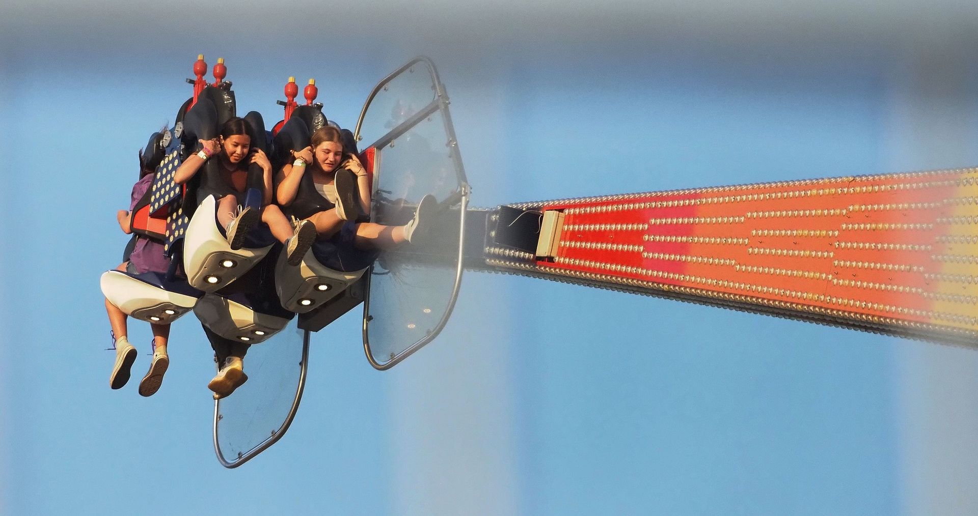 Two people in seats are suspended in mid-air on a rotating thrill ride, positioned against a clear blue sky.