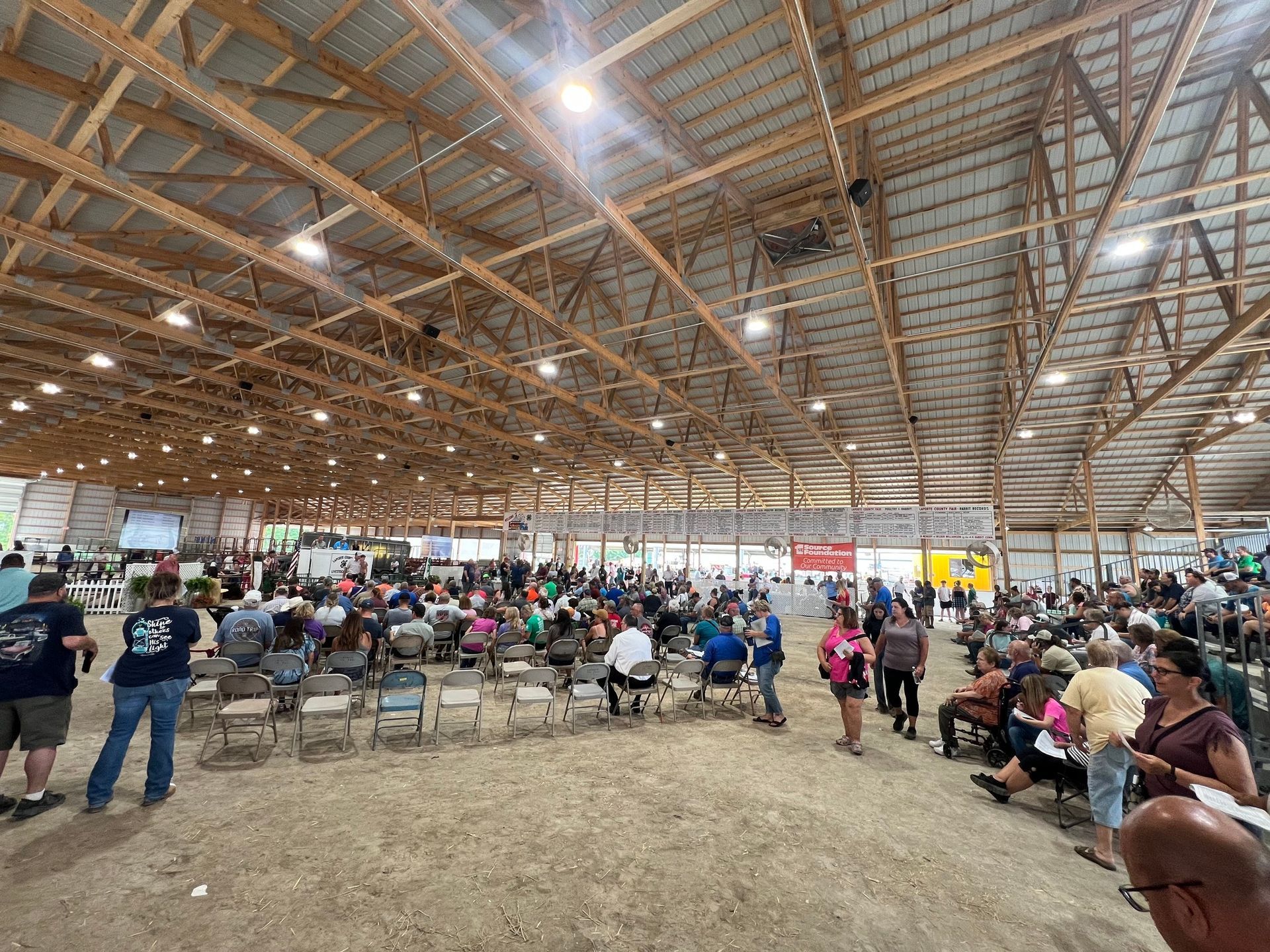 A large indoor agricultural pavilion filled with rows of chairs and a crowd of people attending an event.