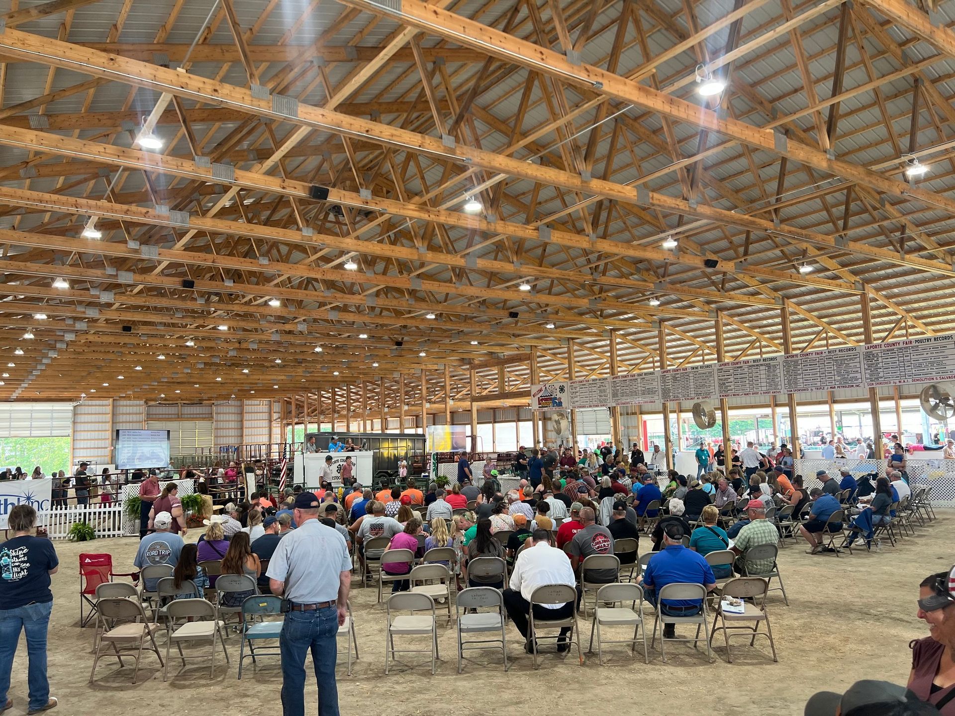 A group of people sit in chairs inside a large, open-air wooden barn structure, facing a presentation screen.