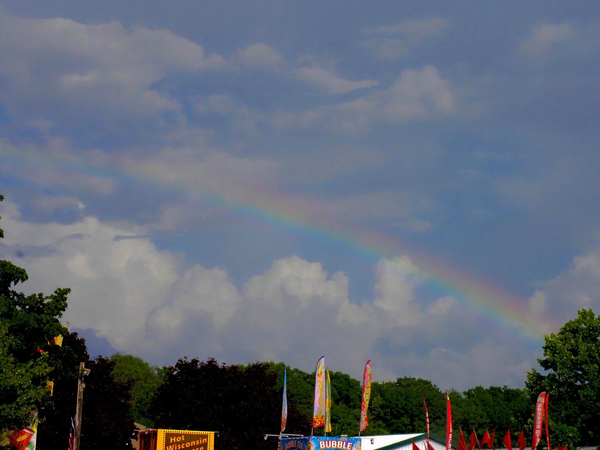 A faint rainbow arches across a blue, cloudy sky above a line of colorful festival flags and dark green trees.