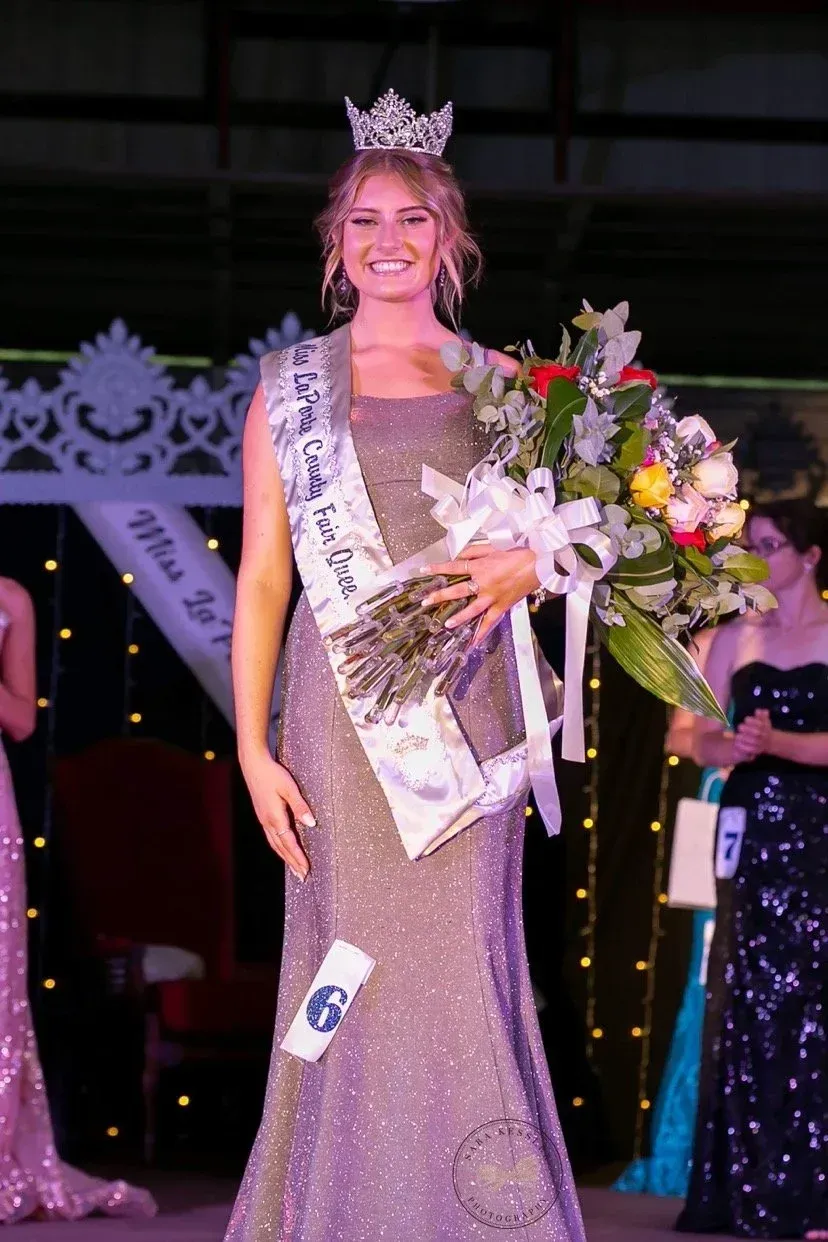 A smiling pageant winner in a sparkling gown and crown holds a bouquet of flowers on stage.