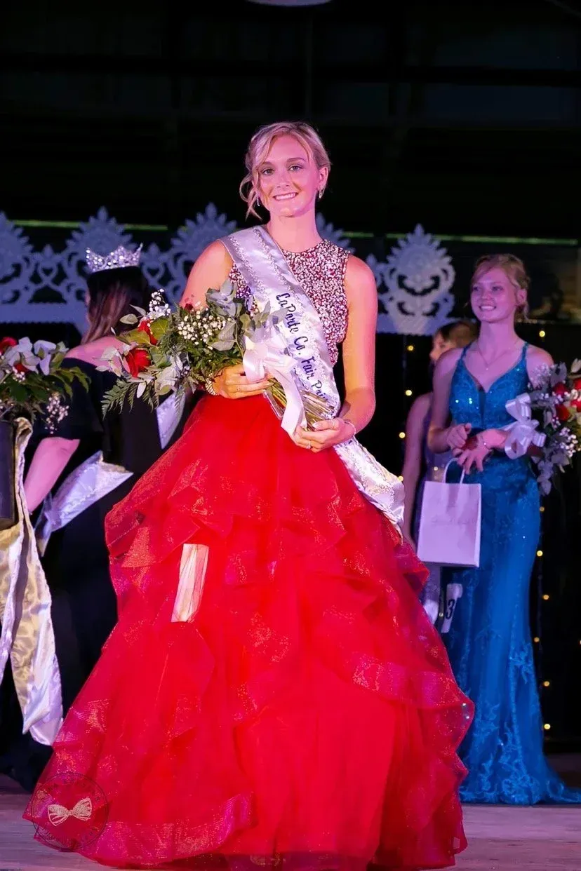 A smiling contestant in a red tiered gown and sash stands on stage holding a bouquet, with others in the background.