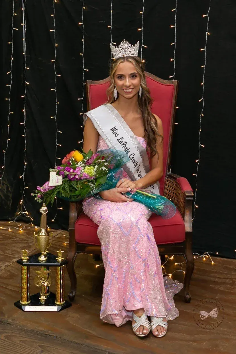 A person wearing a crown and a pageant sash sits in a red chair holding a bouquet of flowers next to a gold trophy.