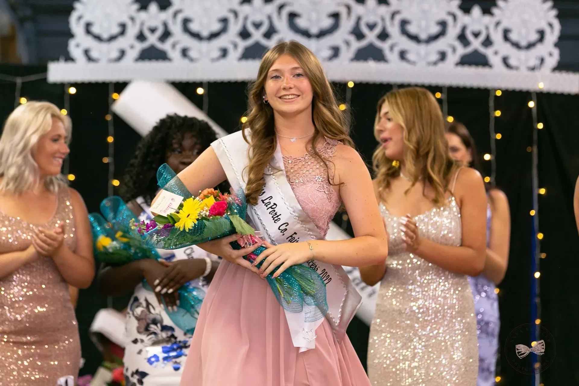 A group of people in formal attire and sashes stand on a decorated stage with string lights, smiling and holding flowers.