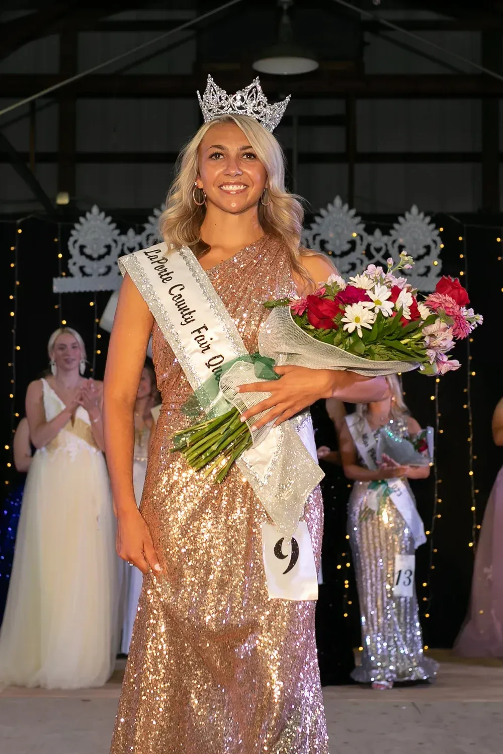 A contestant wearing a sparkly gold dress and crown holds a flower bouquet on a pageant stage, number 9 pinned to sash.