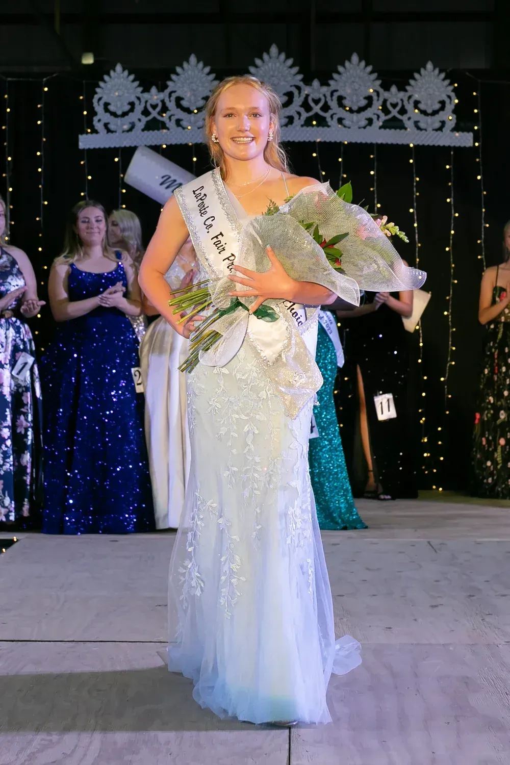 A pageant contestant in a light blue, beaded floor-length gown holds flowers and wears a sash on a stage with lights.