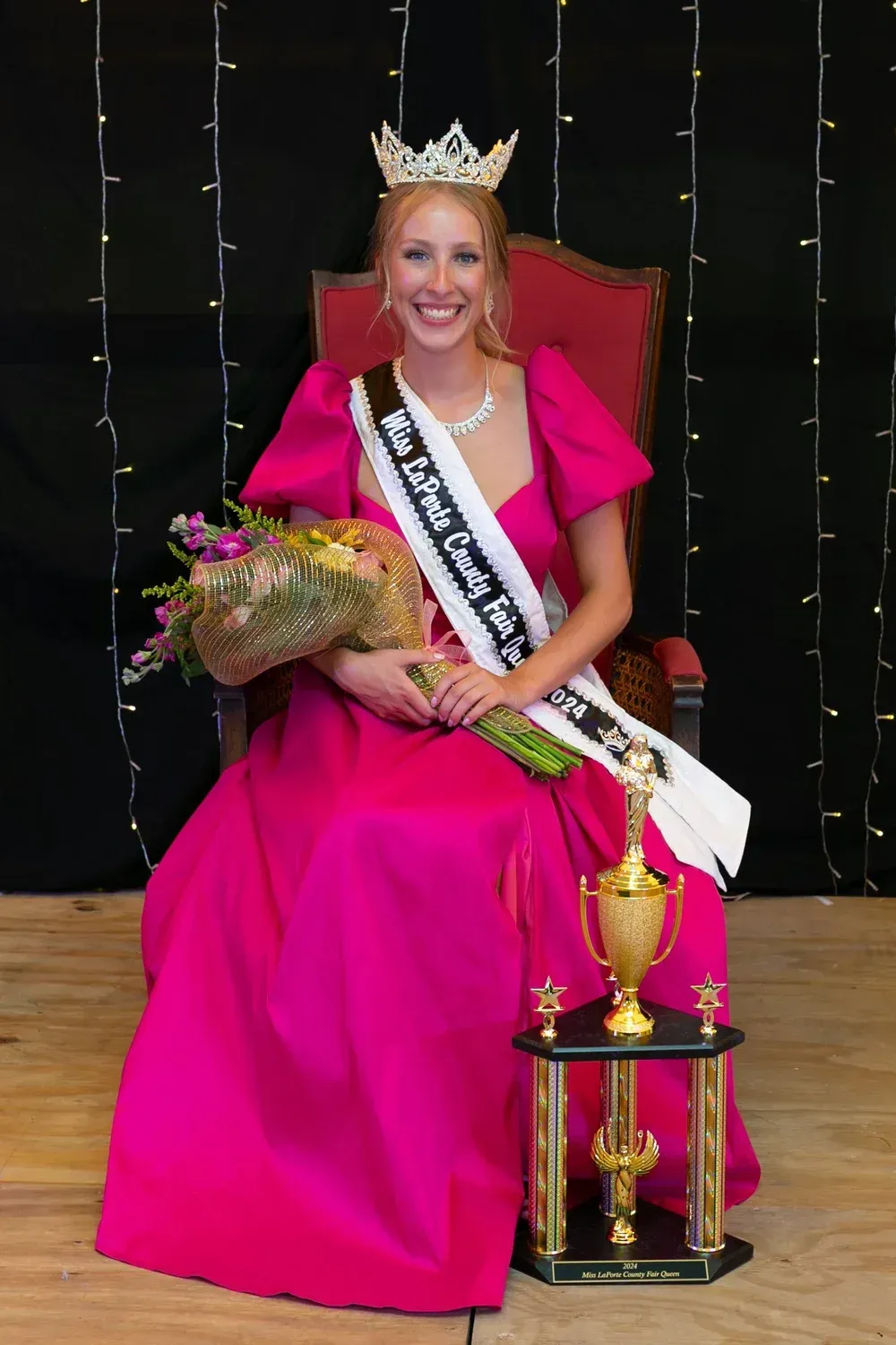 A pageant queen in a vibrant pink gown, wearing a crown and sash, sits holding a bouquet with a trophy at her side.