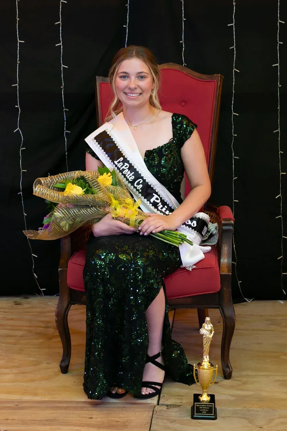 A person wearing a sequined green gown and a pageant sash sits in a red chair holding yellow flowers next to a trophy.