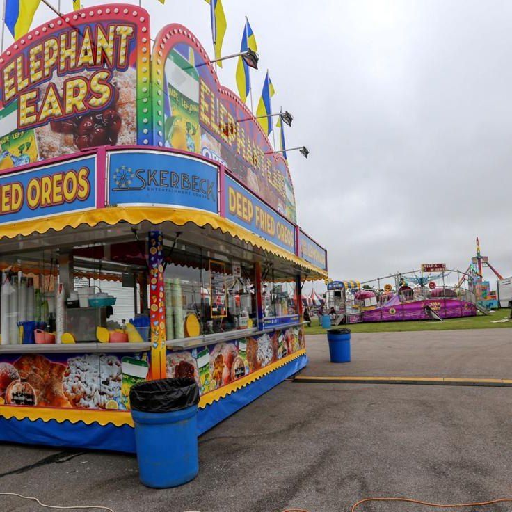 A colorful fair food stand advertising elephant ears and deep-fried Oreos, with amusement rides visible in the distance.