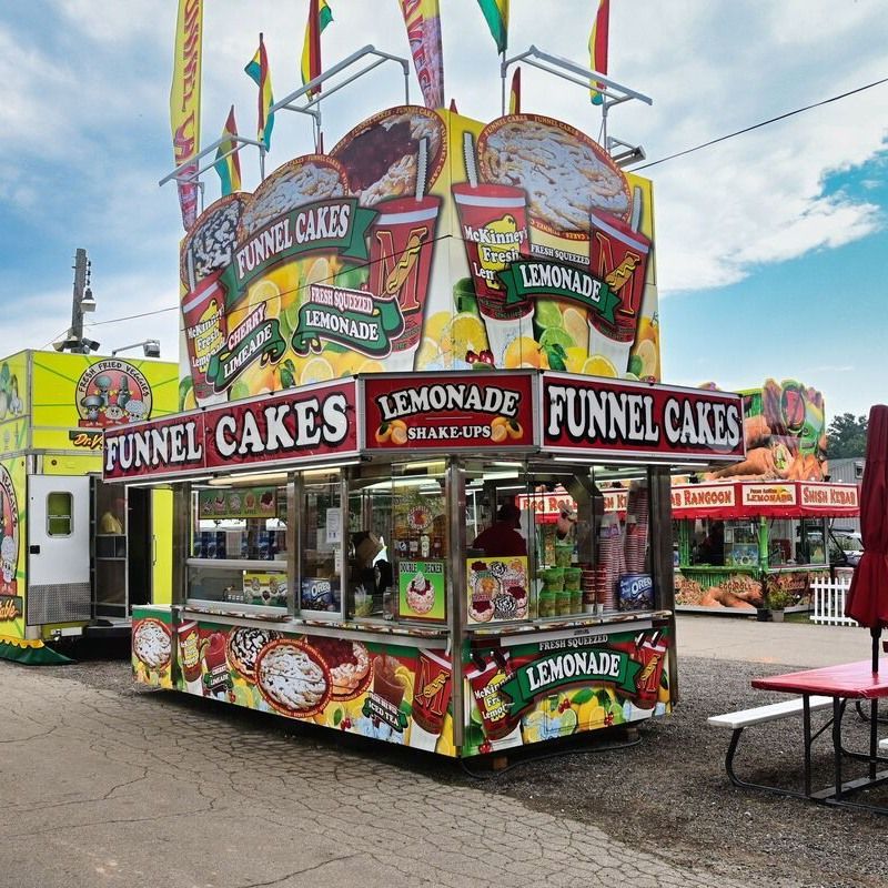 A colorful, rectangular fair food stand selling funnel cakes and lemonade, situated outdoors on a gravel lot.