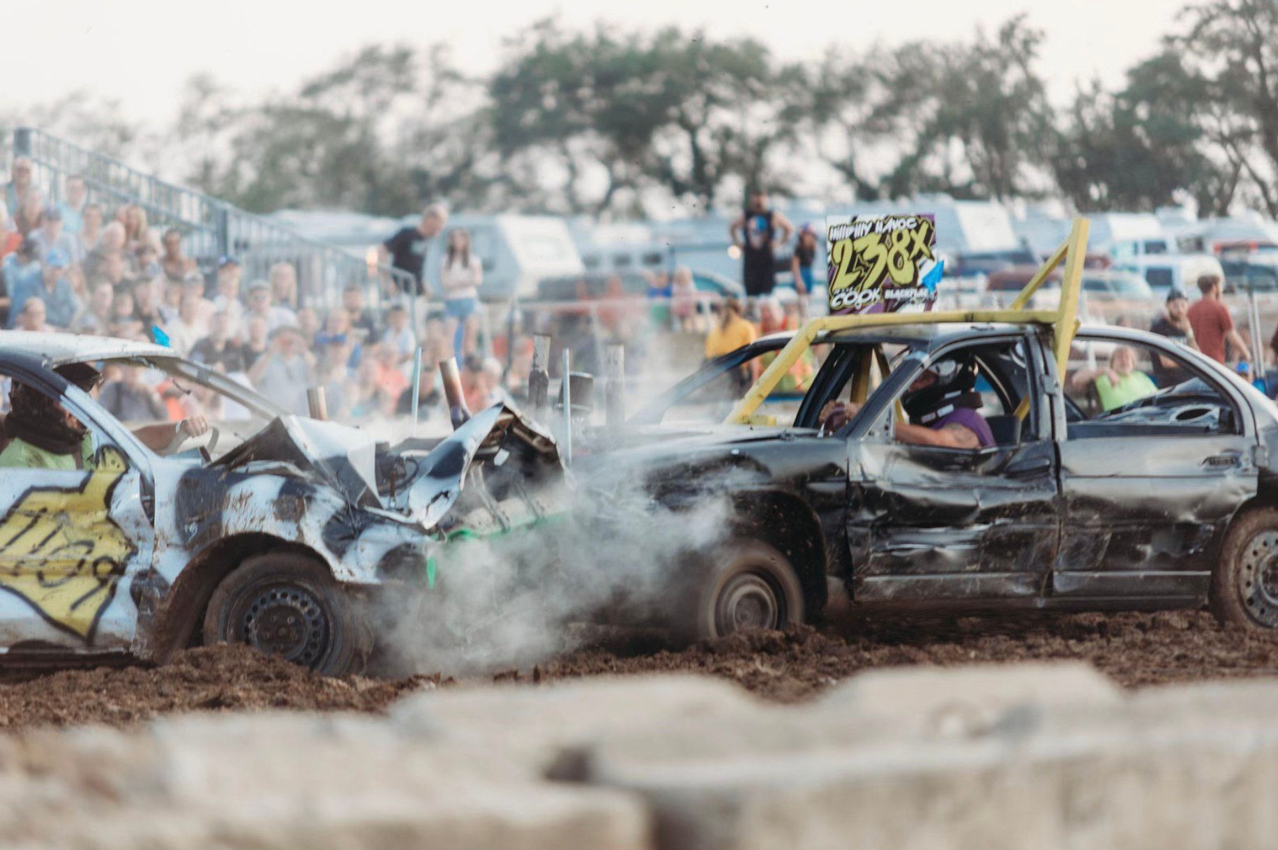 Two cars collide in a muddy demolition derby as a crowd watches from behind a fence.