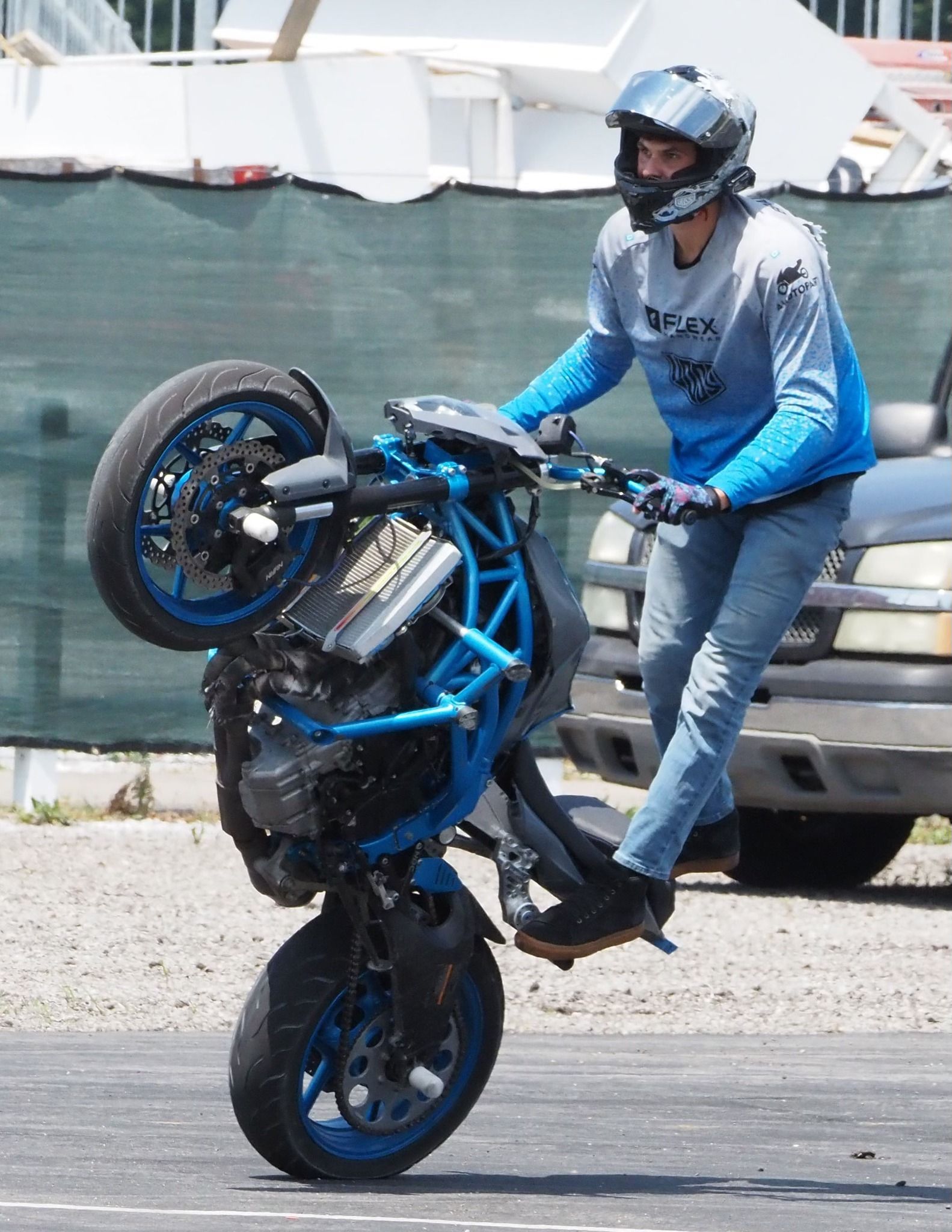 A person in a blue and gray long-sleeved shirt and helmet riding a blue motorcycle on its back wheel in a parking lot.