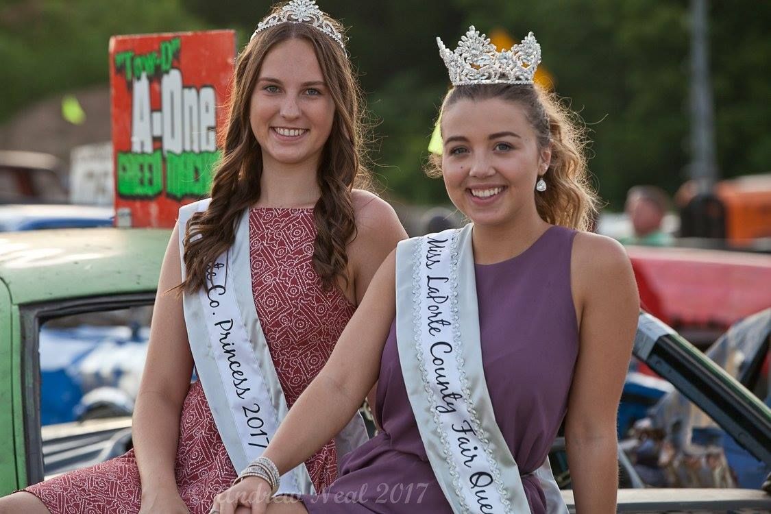Two pageant queens wearing crowns and sashes pose smiling in front of a car at an outdoor event.