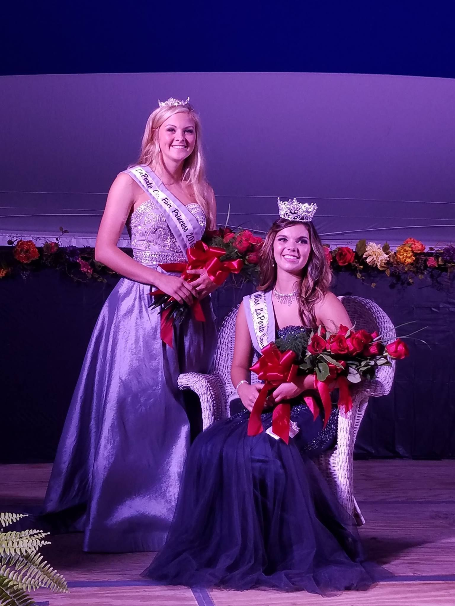Two pageant participants in gowns with sashes and crowns, one standing and one seated in a chair, holding bouquets.
