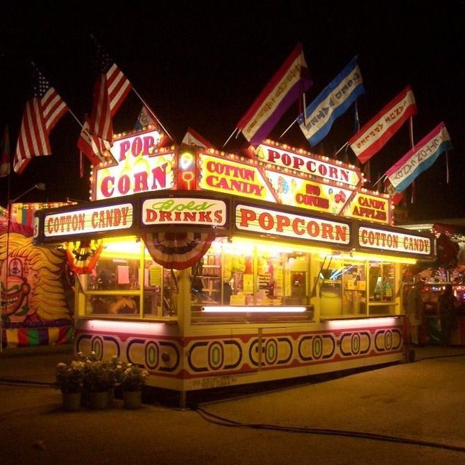 A brightly lit carnival food stand at night, advertising popcorn, cotton candy, and cold drinks with festive flags.